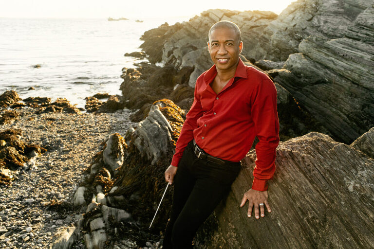 Nicolás Dosman | Director of Choral Studies at the University of Southern Maine Osher School of Music. Leaning on the rocks of the Maine Coast with baton.