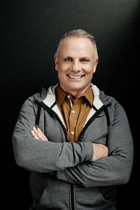 Mike Throne, leadership coach,  standing on a black background in a tan button shirt and gray hoodie with his arms crossed.