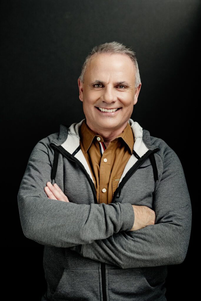 Mike Throne, leadership coach,  standing on a black background in a tan button shirt and gray hoodie with his arms crossed.