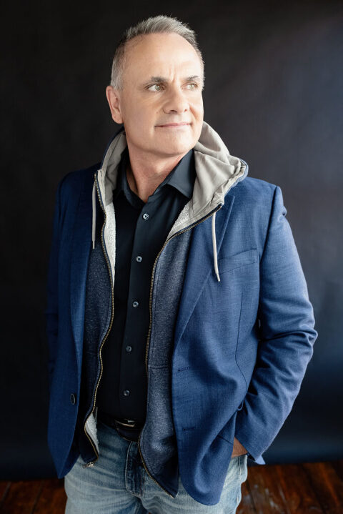 Mike Thorne, Leadership Coach, Standing on black background in black button down shirt with hoodie as mid layer and blue blazer on top. He is looking off towards the light on the right.