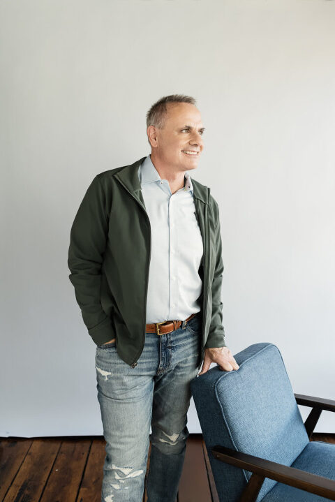 Mike Thorne, Leadership Coach, standing in tattered jeans, light blue button down and unzipped green jacket. He is leaning on a blue chair.