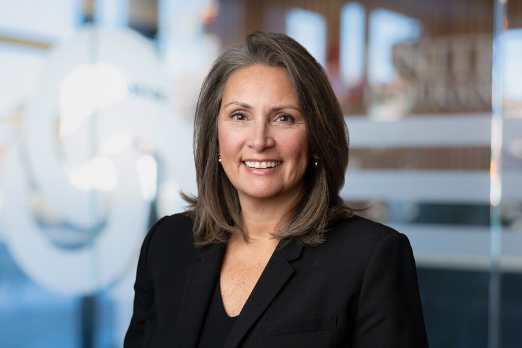 Headshot of a woman wearing a black blazer and shirt for United Insurance. Background has been replaced with glass door with logo on it blurred. Taken by Wohler & Co. Digital Background Change for Headshots
