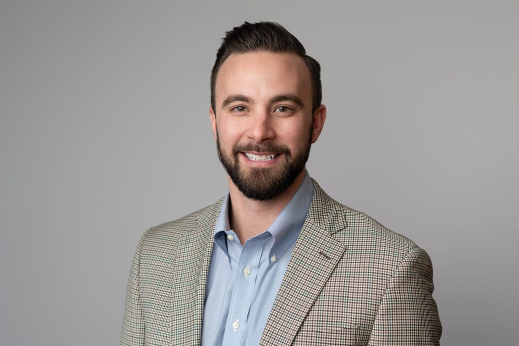 Headshot of a man wearing blue button up shirt with patterned tan blazer for United Insurance. Background has been replaced with glass door with logo on it blurred. Taken by Wohler & Co. Digital Background Change for Headshots