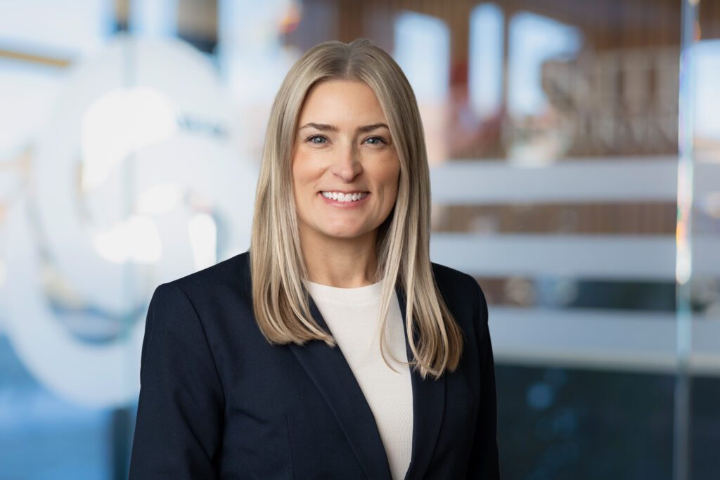 Headshot of a woman with blonde hair wearing a cream shirt with navy blazer for United Insurance. Background has been replaced with glass door with logo on it blurred Taken by Wohler & Co. with Digital Background Change for Headshots.